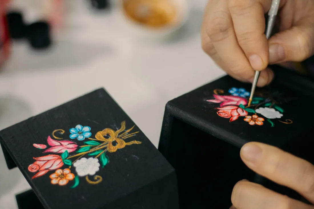Mano con un pincel fino trazando un motivo floral sobre una caja lacada de fondo negro